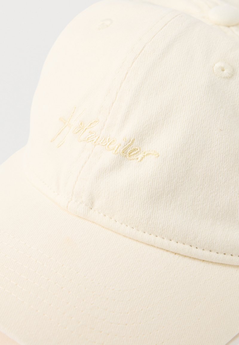 Gebroken witte katoenen honkbalpet met klein geborduurd "H:Traveller"-logo op het voorpaneel, zichtbare stiksel details en een gebogen klep.