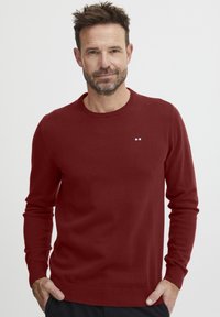 Pulover burgund dintr-un material moale, cu guler rotund, manșete și tiv cu dungi, având un mic logo cu dungi roșii, albe și albastre pe piept.