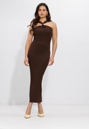 Vrouw met lang donker haar, gekleed in een mouwloze, donkerbruine, aansluitende midi-jurk en crème hakken, staand tegen een effen witte achtergrond.