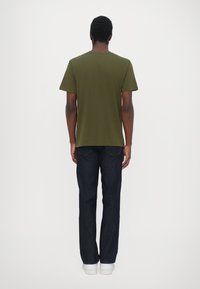 Tricou din bumbac olive cu mânecă scurtă, purtat cu blugi negri și adidași albi, văzut din spate.