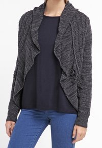 Grau strukturierte Strickjacke mit Schalkragen, mit Fransenakzenten und offenem Vorderteil. Getragen über einem dunklen Oberteil und kombiniert mit blauen Jeans.