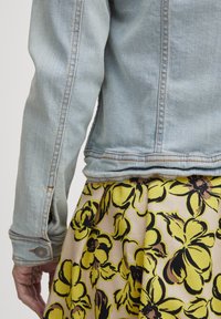 Šviesiai mėlynas džinsinis švarkas su matomomis siūlėmis, ilgomis rankovėmis ir mygtukų uždarymu ant rankogalių, derinamas su geltonu sijonu, puoštu gėlėmis.