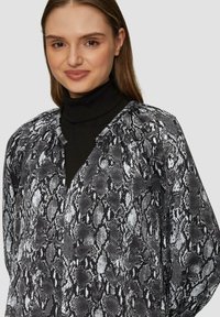 Blouse imprimé serpent noir et blanc avec une coupe décontractée, un décolleté en V et une texture douce. Portée par-dessus un col roulé noir pour un style en couches.