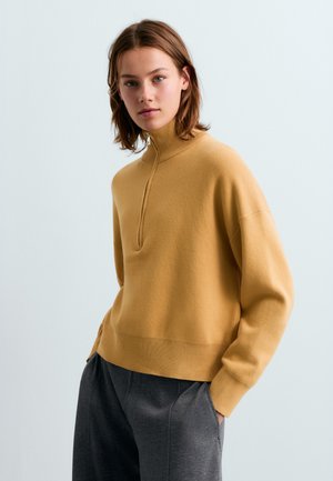 Jonge vrouw met kort bruin haar, dragend een mosterdgele trui met rits en donkergrijze broek, staand met handen in de zakken.