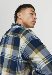 Camisa de franela con un patrón de cuadros multicolor en azul marino, verde y amarillo, con cuello tipo solapa y cierre de botones en la parte delantera.