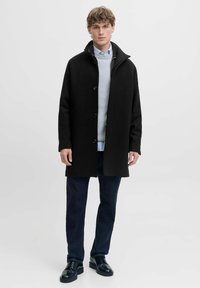 Must tokk, millel on kõrge krae, esipaneelil nööbid, kantud halli sviitri ja helesinise särk üle, koos tumedate pükste ja mustade kingadega.