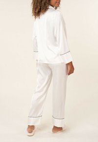 Ensemble de pyjama en satin blanc comprenant un haut à boutons à manches longues avec des bordures noires et un pantalon ample, texture lisse et silhouette décontractée.