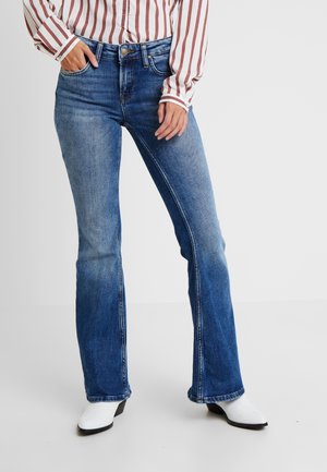Femme portant un jean bootcut bleu, des bottes blanches et une chemise blanche à manches longues avec des rayures rouges verticales, debout avec une main dans la poche.