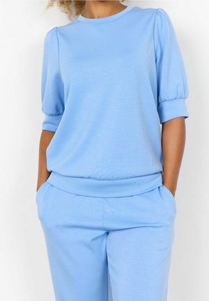 Frau trägt hellblaues kurzärmeliges Oberteil mit aufgeschlagenen Puffärmeln und dazu passende Hose, Hände in den Taschen, vor schlichtem weißem Hintergrund.