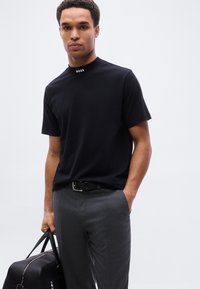 Άνδρας που φοράει μαύρο T-shirt BOSS και γκρι παντελόνι, κρατώντας με το ένα χέρι μαύρη δερμάτινη τσάντα duffel, ενώ το άλλο χέρι είναι στην τσέπη, στέκεται μπροστά σε λευκό φόντο.