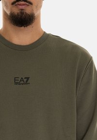 Maglietta a maniche lunghe verde oliva realizzata in tessuto morbido, con scollo a girocollo e logo EA7 in nero. Design semplice e aderente.