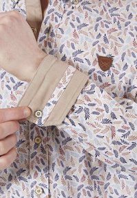 Chemise à manches longues avec un motif de feuilles blanc et multicolore, col beige, détails de boutons et un petit patch logo marron sur la poitrine.
