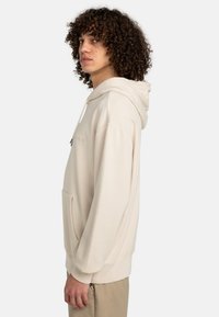 Sudadera con capucha beige con un bolsillo tipo canguro en la parte frontal, puños acanalados y textura suave. Presenta una marca discreta en un color contrastante.