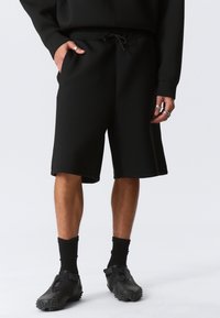 Schwarze Shorts mit lockerer Passform, Kordelzugbund und seitlichen Taschen. Aus weichem Stoff mit glatter Textur gefertigt. Dazu schwarze Schuhe.