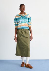 Fleecegenser med turkiser, oransje og hvite striper; olivengrønn skjørt; brune sneakers med gule detaljer; hvite ribbestrikkede sokker.