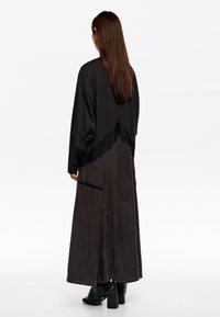 Długa czarna sukienka z textured materiału, z koronkowym akcentem w talii i prostym krojem. Modelka ma na sobie czarne botki na obcasie.