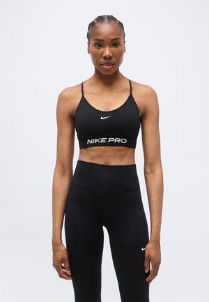 Naine, kes kannab musta Nike Pro spordirinnahoidjat ja kõrge vöökohaga musti retuuse, seisab sirgelt halli tausta ees.