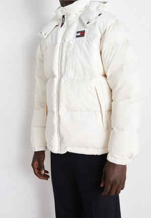 Homme portant une doudoune blanche avec une poche zippée sur la poitrine et le logo Tommy Jeans, associée à un pantalon noir sur fond blanc.