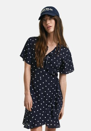Marineblauwe polkadot wikkeljurk met korte mouwen en een rucheszoom. Gecombineerd met een marineblauwe pet met witte tekst aan de voorkant.