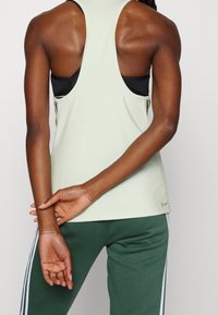 Débardeur de sport vert clair sans manches, texture lisse, conception dos nageur, porté sur un soutien-gorge de sport noir, accompagné d'un pantalon vert foncé.