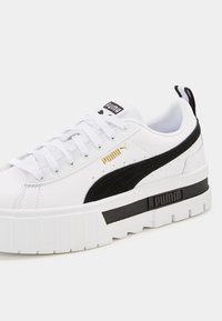 Sneaker bianco con suola spessa, accenti in suede nera e logo dorato. Presenta lacci bianchi, superficie testurizzata e un'etichetta sul tallone con il marchio.