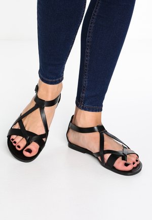 Jalkineet, joissa on mustat pintaletkut ja nilkkaremmit, yhdistettynä tummansinisiin, tiukkoihin farkkuihin valkoista taustaa vasten.