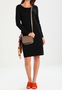 Robe noire à manches longues, design ajusté. Sac en bandoulière marron matelassé avec une sangle d'épaule. Talons ouverts orange vif. Écharpe à motifs attachée au sac.
