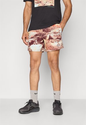 Szorty Nike z wielokolorowym, abstrakcyjnym wzorem w odcieniach brązu, kremu i pomarańczowego, w połączeniu z szarymi skarpetami Nike i czarnymi butami.