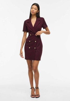 Kobieta stojąca, ubrana w bordową sukienkę z krótkim rękawem, z podwójnym rzędem złotych guzików, oraz czarne sandały na pasku przy kostce, na białym tle.