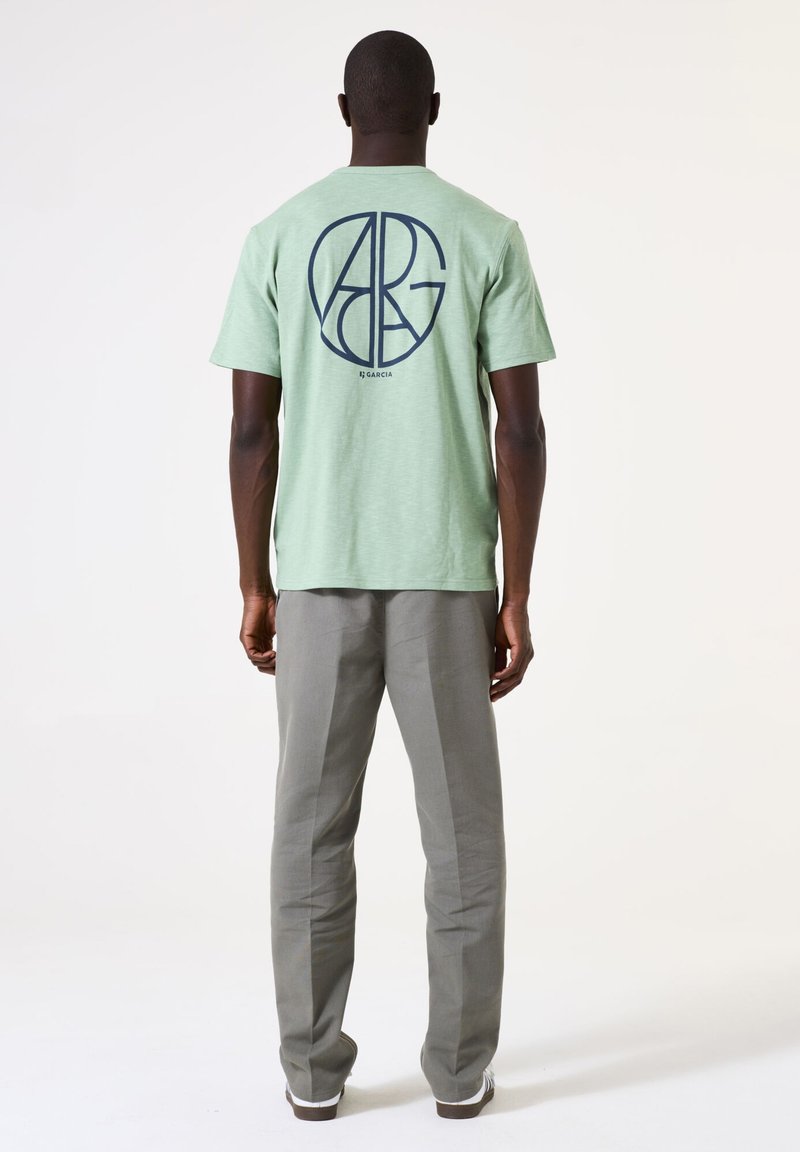 Groen katoenen T-shirt met een marineblauw logo ontwerp op de achterkant, gecombineerd met lichtgrijze broeken. Eenvoudig en recht gesneden ontwerp.