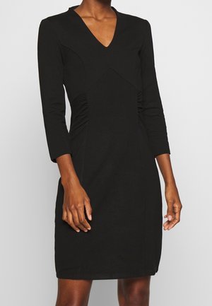 Woman wearing a fitted black dress with three-quarter sleeves and a V-neckline against a plain light gray background.