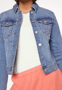 Jeansjacka i ljusblått, med silvriga tryckknappar, två bröstfickor och en kroppsnära design. Tillsammans med en vit topp och en korallfärgad kjol.