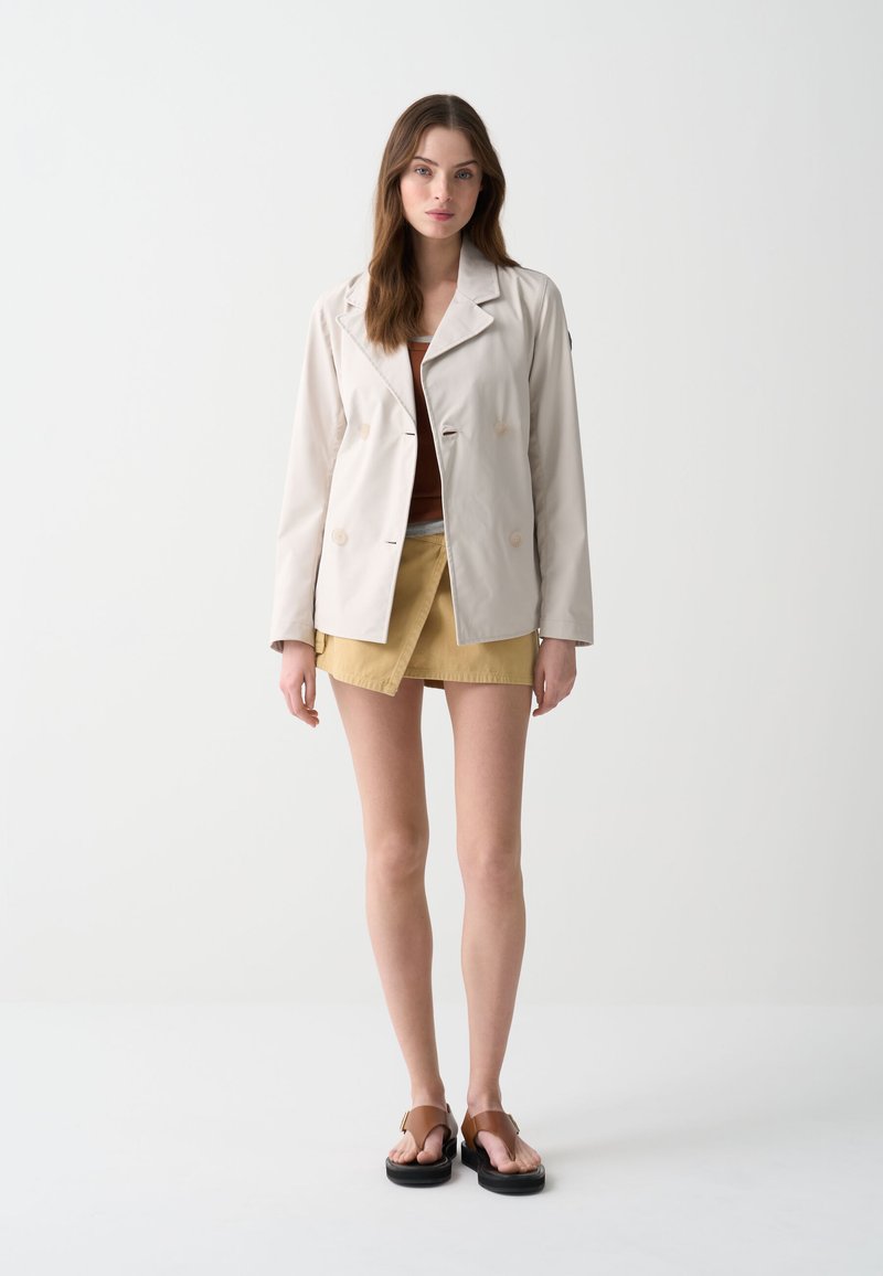 Veste beige à double boutonnage, en tissu léger, avec revers à notch, associée à un short-jupe moutarde et des sandales marron. Palette de couleurs neutres.