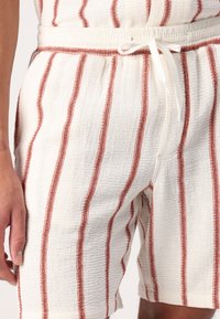 Shorts blancos de tejido entrelazado con rayas rojas horizontales, cintura con cordón y tela texturizada. Detalle en primer plano que muestra las costuras y el patrón.