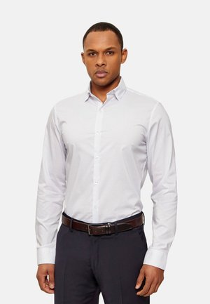 Hombre vestido con una camisa blanca de vestir ajustada con estampado, pantalones oscuros y un cinturón marrón, de pie contra un fondo claro y liso.