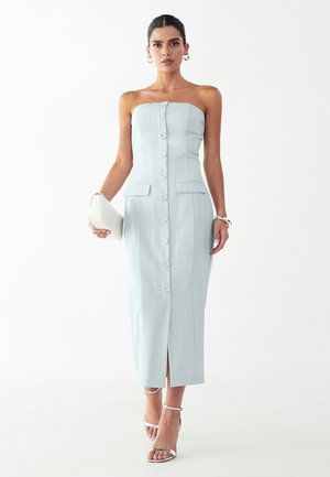 Kobieta w jasnoniebieskiej, bezrękawnikowej sukience midi z guzikami z przodu, trzymająca białą kopertówkę i mająca na sobie białe szpilki.