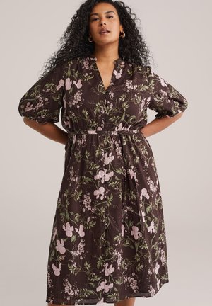 Žena s kučeravými vlasmi, nosiaca zlaté kruhové náušnice a tmavohnedé kvetinové šaty s krátkymi nafúknutými rukávmi a vôľou v páse.