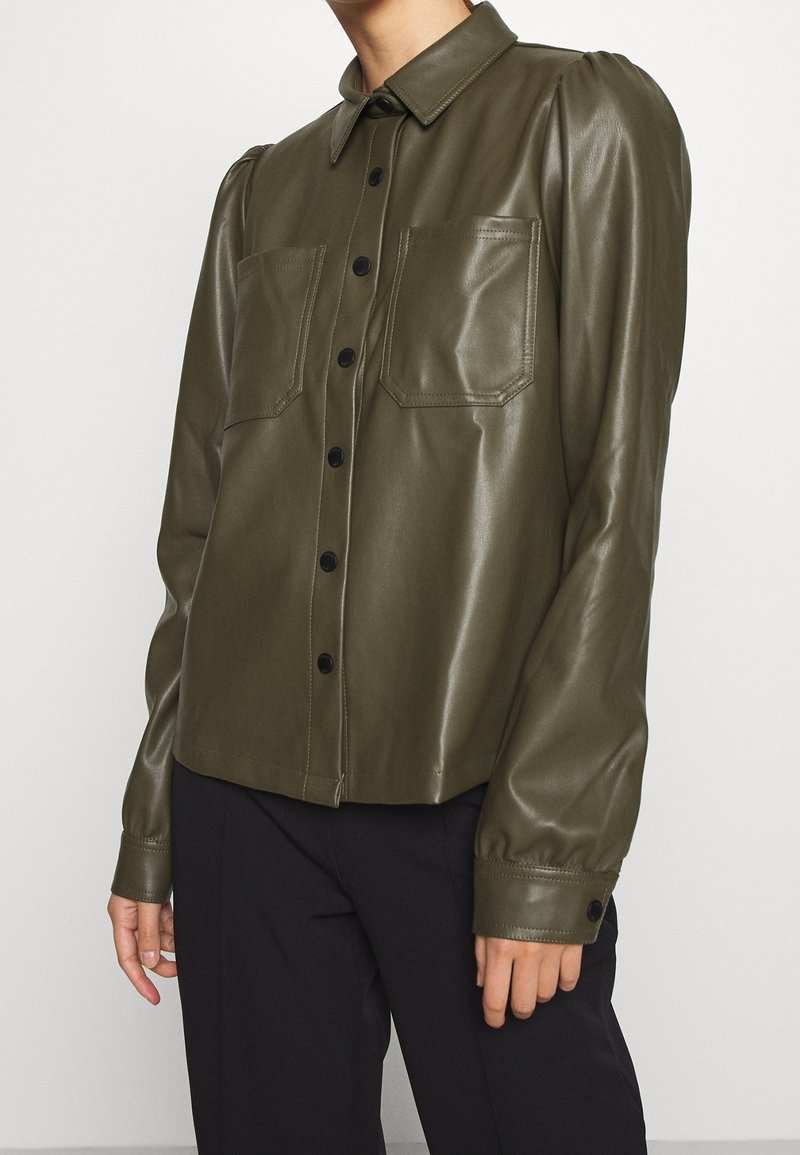 Camicia in ecopelle verde oliva con colletto, chiusura frontale a bottoni, due tasche sul petto e maniche arricciate. Tessuto liscio, dettagli con bottoni.