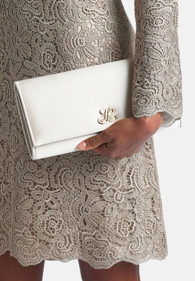 Main tenant une pochette blanche avec fermoir doré, portant une robe beige en dentelle avec motifs floraux et bords festonnés.