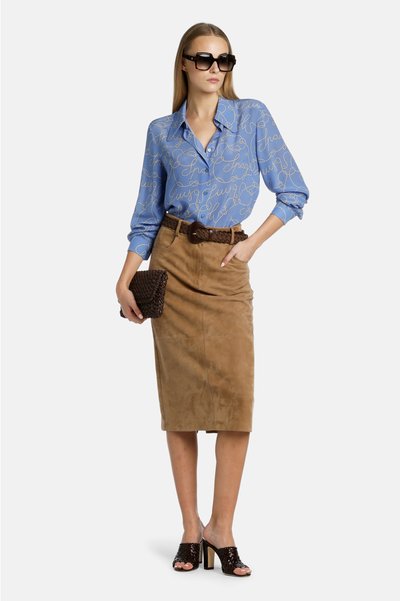 Blauwe overhemd met knopen en een gouden schriftontwerp, gecombineerd met een beige suède kokerrok. Zwarte geweven clutch en gespilde muiltjes met een blokhak.