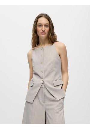 Mujer con cabello largo y ondulado que lleva un chaleco beige sin mangas con botones y pantalones anchos a juego, manos en los bolsillos, fondo neutro.