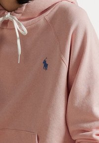 Detailní záběr na osobu v růžové mikině s bílými šňůrkami a malým námořnickým logem jezdce na polo na hrudi.