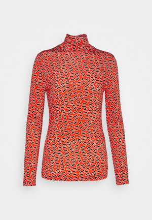 Femme portant un jean noir taille haute, des escarpins noirs pointus, un haut à manches longues orange à motifs et un sac à épaule noir devant un mur uni.