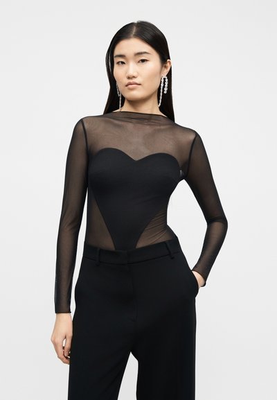 Γυναίκα με μακριά σκούρα μαλλιά φοράει μαύρο διαφανές μπλουζάκι με μακριά μανίκια και το μπούστο σε σχήμα καρδιάς, μαζί με μαύρο παντελόνι ψηλόμεσο. Έχει το ένα χέρι στην τσέπη.