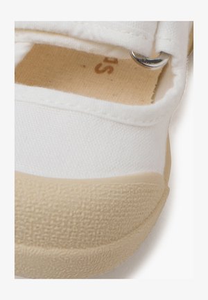 Baskets en toile blanches avec une semelle en caoutchouc beige, dotées d'une sangle velcro et d'un accent en métal argenté. Tissu texturé avec des détails cousus.