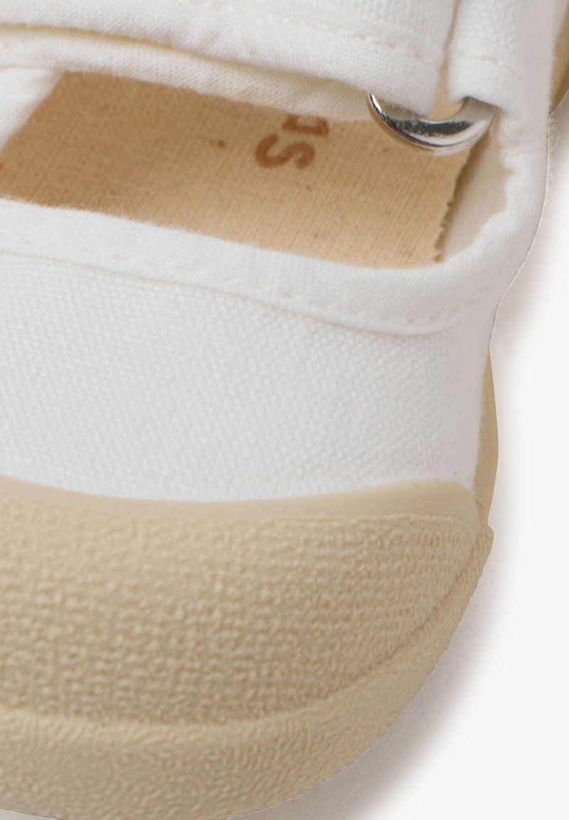Zapatilla de lona blanca con suela de goma beige, que presenta una correa de gancho y bucle y un acento de metal plateado. Tejido texturizado con detalles cosidos.