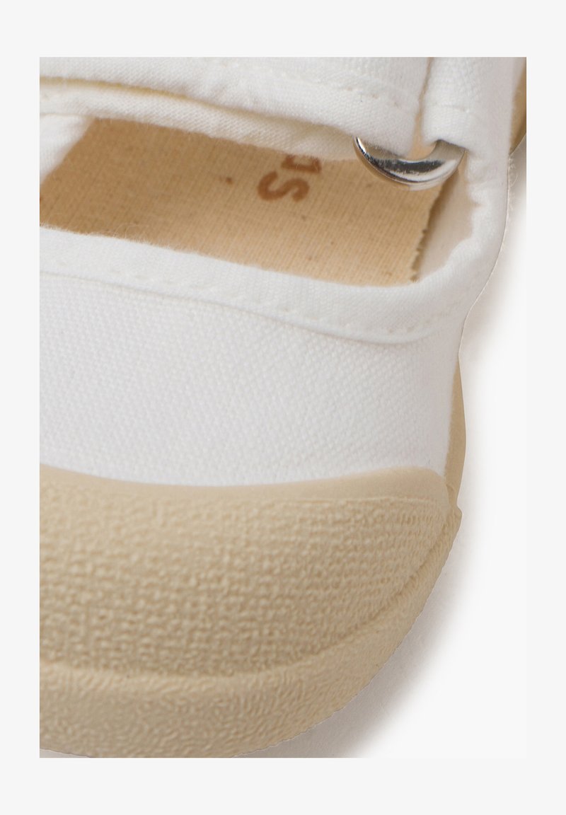 Bílé plátěné boty s béžovou gumovou podešví, mají suchý zip a stříbrný kovový akcent. Texturovaná tkanina s přišitými detaily.