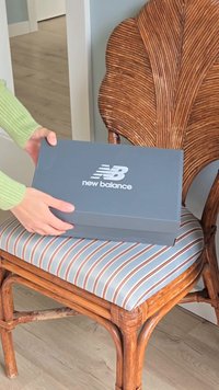 Mani che posizionano una scatola di scarpe grigia New Balance su una sedia in legno imbottita a strisce con uno schienale decorativo.