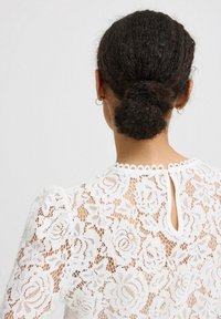 Blusa bianca in pizzo con motivo floreale, chiusura con bottoni sul retro, scollatura scallop e maniche a sbuffo. Tessuto in pizzo testurizzato visibile.