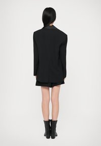 Zwarte tailored blazer met een gestructureerd ontwerp, vergezeld van zwarte shorts en enkellaarsjes, tegen een effen witte achtergrond.